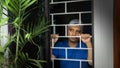 Latin man with blue shirt locked in his house looking out through the window, leaning against security bars