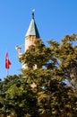 Close-up view minaret which is covered by green tree leaves. Blue sky background. Turkish flag near the minaret