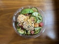 Close up view of a plastic bowl of salmon and tuna poke with crab salad, cucumbers, and avocado on a wooden table