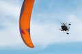 A close-up view of a powered paraglider mid-flight shows the orange wing and pilot s engine under a bright blue sky, capturing Royalty Free Stock Photo