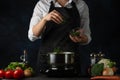 Close-up view of the professional chef in black apron adding chopped parsley in boiling water for soup on dark blue background. Royalty Free Stock Photo