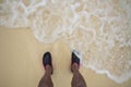 Close up view of rolling waves on men`s feet standing on white sand beach in swimming shoes Royalty Free Stock Photo