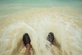 Close up view of rolling waves on men`s feet standing on white sand beach in swimming shoes Royalty Free Stock Photo