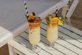 Close-up view of two glasses of cocktail on white table on sandy beach under sun lounger.