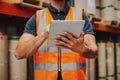 Close-up of warehouse worker in orange vest holding digital tablet working while checking on inventory and order