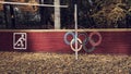 Close up on white bicycle sign or icon and colored sign of the Olympic rings on a brown wooden fence in the public park.