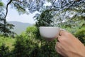 Close up white coffee cup is held by hands of traveler against beautiful green nature and mountain background. Royalty Free Stock Photo