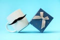 Close up of white mug with a felt moustache and present box on a blue background. Concept of Father's day