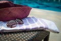 Close-up of white and purple Turkish towel, sunglasses and straw hat on rattan lounger with blue swimming pool as background. Royalty Free Stock Photo