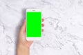 Close up woman hand holding white mobile phone with blank green screen on white marble background, front view with copy space. Royalty Free Stock Photo