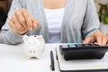 Close up of woman hand putting coin in piggy bank while using calculator on white desk at home. Family expense  savings  collect Royalty Free Stock Photo