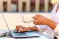 Close up of woman hands holding a cup of coffee and typing on laptop outdoors at cafe table Royalty Free Stock Photo