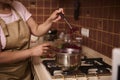 Close-up of a woman, a housewife in an apron, standing at the kitchen stove and making cherry berry jam in metal saucepan in the Royalty Free Stock Photo