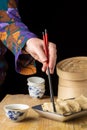 Close-up of woman`s hand with chopsticks holding a gyoza on plate, on wooden table with soy bowl and steamer, Royalty Free Stock Photo