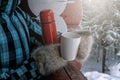 Close-up. Woman in a warm plaid jacket holds a red thermos with a hot drink and a white mug. Winter forest background Royalty Free Stock Photo