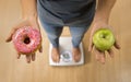 Close up woman on weight scale holding in her hand apple fruit and donut as choice of healthy versus unhealthy food Royalty Free Stock Photo