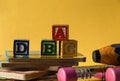 Closeup of Woodblock letters a, b, c, d, children reading books, big yellow and blue pencils in front of a yellow background.