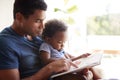 Close up of young adult African American father reading a book with his two year old son, close up, side view, backlit