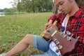 Close up young asian man in red shirt leaning against a tree and playing acoustic guitar in beautiful outdoor park. Shallow depth Royalty Free Stock Photo