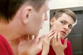 Close up of young attractive man with problematic skin and scars from acne looks in the mirror in the bathroom