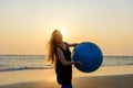 Close-up of young beautiful woman with long blonde hair holds big fitness ball standing on beach in the light of setting sun