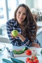 Close up of young brunette woman smiling with apple in her hand chopping green vegetables at home in the kitchen Royalty Free Stock Photo