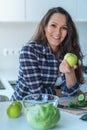 Close up of young brunette woman smiling with apple in her hand chopping green vegetables at home in the kitchen Royalty Free Stock Photo