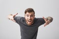 Close up of young cheerful bearded tattooed good-looking guy with stylish hairstyle in casual gray shirt spreading hands