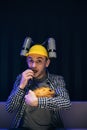 Close up of the young funny man with beer helmet on the head eating chips while watching movie on the sofa in the living Royalty Free Stock Photo