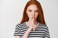 Close-up of young ginger girl with serious face showing taboo sign, hushing at you, telling a secret, white background Royalty Free Stock Photo