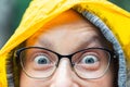 Close-up young happy woman portrait with wide opened amazed eyes wearing glasses with raindrops and bright yellow raincoat hood