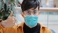 Close-up young indian guy in medical mask posing indoors looking at camera showing thumbs up sign approval agreement Royalty Free Stock Photo