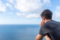 Close-up of young man on his back leaning against a wooden railing, looking at a sunny seascape with white clouds