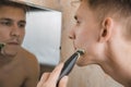 Close up of young man looking to mirror and shaving beard with trimmer or electric shaver Royalty Free Stock Photo