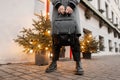 Close-up of a young man standing near the building in a stylish plaid coat in vintage trousers in fashionable boots with a leather