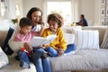Close up of young mother sitting on a sofa in the living room reading a book to her two children, father sitting at a table in the