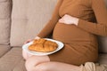 Close up of young pregnant woman enjoys eating croissant on the sofa. Unhealthy pastry during pregnancy concept