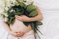 Close up of young pregnant woman hands touching gently tummy lying on comfortable bed holding beautiful fresh summer flowers. Royalty Free Stock Photo