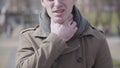 Close-up of young sick man couching and rubbing throat. Portrait of ill Caucasian guy outdoors on sunny autumn or spring