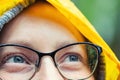 Close-up young tired happy woman portrait wearing glasses with raindrops and bright yellow raincoat hood during rain outdoors