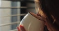 Close-up young woman drinking hot coffee or tea standing near window with blinds at sunrise and looking out of window Royalty Free Stock Photo