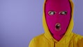 Close up of young woman in pink balaclava and yellow hood grimacing face on purple background. Crazy female in mask