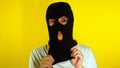 Close up young woman puts on black balaclava on yellow background. Secretive female puts on mask, looking at camera.