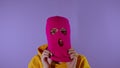 Close up young woman puts on pink balaclava on purple background. Secretive female puts on mask, looking at camera.