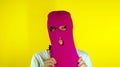 Close up young woman puts on pink balaclava on yellow background. Secretive female puts on mask, looking at camera.