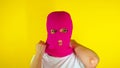 Close up young woman puts on pink balaclava on yellow background. Secretive female puts on mask, looking at camera.