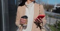 Close up young woman using smartphone and drinking coffee to go outdoors, walking down the city street. Royalty Free Stock Photo