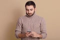 Close upportrait of handsome young man using his smartphone while standing isolated over beige background, looking at his device