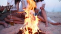 Close view of friends frying sausages sitting around bonfire, drinking beer, playing guitar on sandy beach. Young group