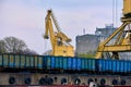 Close view of river port crane loading open-top gondola cars of freight train on cloudy day. Empty river drag boats or